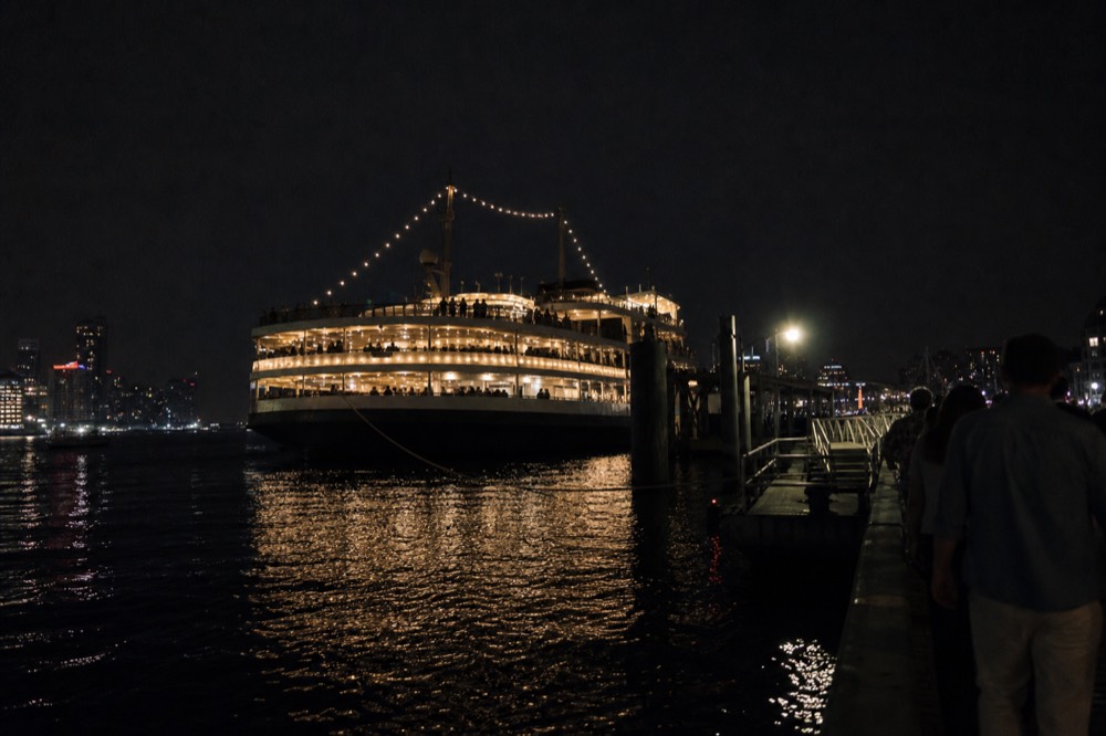 Dinner cruise vessel lit up on dark harbor water with city skyline behind
