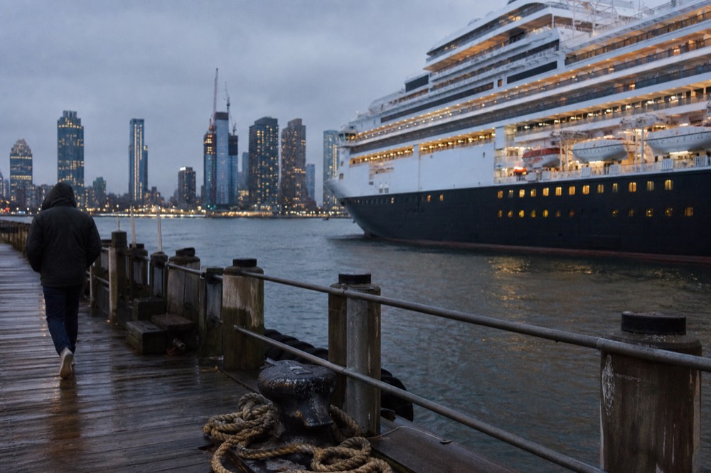 A lit cruise vessel docked at a Seaport pier at dusk with the Boston skyline behind it