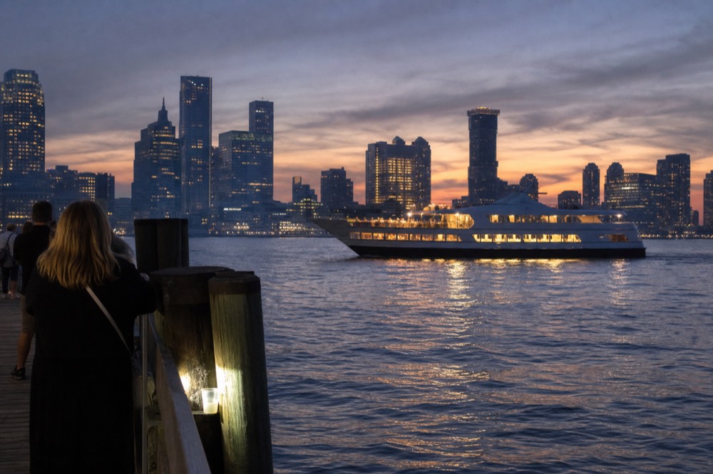 A dinner cruise boat lit up at dusk on Boston Harbor with the skyline behind it