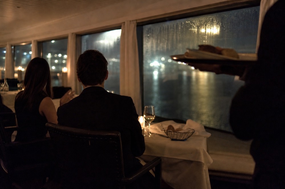 Couples in cocktail attire at a set dinner table aboard a cruise ship, harbor lights visible through windows