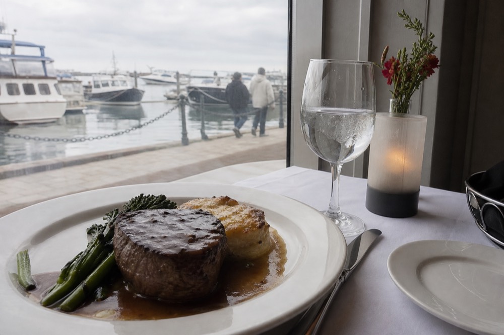 A plated fine-dining entree on a white tablecloth beside a harbor window